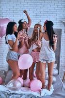 feliz de estar cerca. Longitud total de cuatro atractivas jóvenes mujeres sonrientes en pijamas brindando entre sí mientras tienen una fiesta de pijamas foto