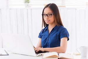 Working on new creative project. Beautiful young Asian woman working on laptop and smiling while sitting at her working place photo