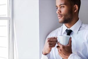 Enjoying his coffee break. Handsome young African man in shirt and tie holding cup with hot drink while looking through the window photo