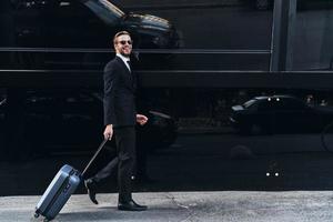 Young and successful. Full length of young man in full suit pulling luggage and smiling while walking outdoors photo