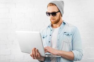 Smart technology for perfect style. Concentrated young bearded man holding laptop and working on it while standing against brick wall photo