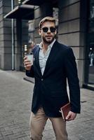 Always in a hurry. Handsome young man in smart casual wear carrying disposable cup while walking through the city street photo