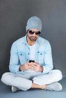 Sending message to friend. Handsome young man in eyewear holding mobile phone while sitting on the floor against grey background photo