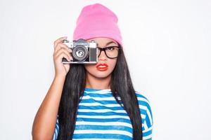 Focusing at you. Attractive young African woman in funky clothes focusing at you with her old-fashioned camera while standing against white background photo