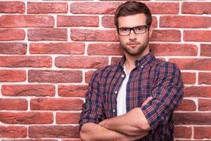 Charming handsome. Handsome young man keeping arms crossed and looking at camera while standing against brick wall photo