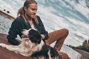 su mejor amigo. un joven apuesto y su perro sentados cerca del lago mientras pasan tiempo al aire libre foto