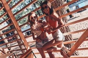 chicas juguetonas. dos atractivas mujeres jóvenes sonriendo y bebiendo cócteles mientras están de pie en el café de la playa foto