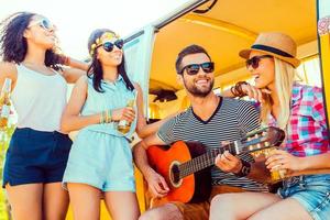 Spending quality time with friends. Handsome young man sitting at minivan and playing guitar while three girls standing close to him and smiling photo