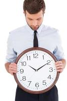 Checking the time. Handsome young man in shirt and tie holding a clock in his hands and looking at it while standing isolated on white background photo