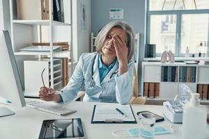 Tired mature female doctor in white lab coat covering her face with hand while sitting in her office photo