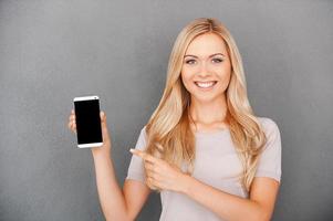 Copy space on her smart phone. Smiling young blond hair woman holding mobile phone and pointing at it while standing against grey background photo
