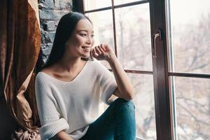 Good memories. Attractive young woman smiling and looking away while sitting on the window sill at home photo