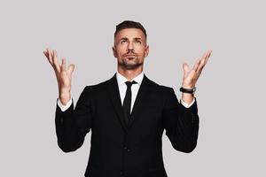 Begging for success. Handsome young man in full suit keeping arms outstretched and looking up while standing against grey background photo