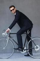 Environmentally friendly transport. Full length of confident young businessman sitting on bicycle and looking at camera while cycling against grey background photo