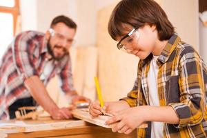 Hardworking is a key to success. Smiling little boymaking measurements on the wooden plank while working with his father in workshop photo