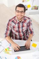 ejecutivo creativo. vista superior de un apuesto joven con anteojos trabajando en una computadora y sonriendo a la cámara mientras se sienta en su lugar de trabajo foto