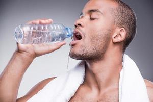 refrescante después del entrenamiento. joven africano sin camisa sosteniendo una botella con agua y sonriendo mientras se enfrenta a un fondo gris foto