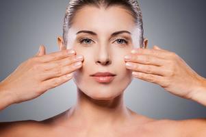 Face care. Beautiful mature woman keeping eyes closed and touching face with hands while standing against grey background photo