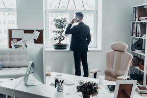 No time for resting. Rear view of young man in full suit talking on the phone and keeping hand in pocket while standing near the window in the office photo