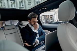 Frustrated young man in full suit working using laptop and covering face with hand while sitting in the car photo