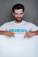 Everything you interested in is here Confident young man in volunteer t-shirt pointing to white board and looking at camera with smile while standing against grey background photo