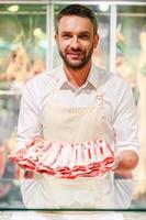 Only the freshest meat Smiling young butcher stretching out slice of meat and looking at camera while standing at supermarket checkout photo