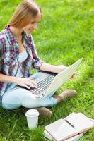 preparándose para los exámenes al aire libre. bella joven estudiante que trabaja en una laptop y sonríe mientras se sienta en un parque con libros a su alrededor foto