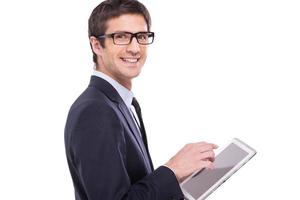 Making your business easier. Side view of cheerful young man in formalwear working on digital tablet and looking at camera while standing isolated on white background photo