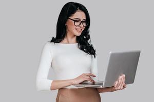 Using laptop. Beautiful young woman in smart casual wear working on laptop while standing against grey background photo