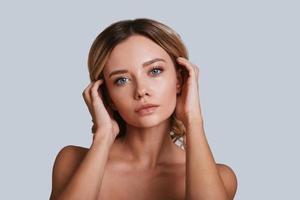 Beautiful perfection. Attractive young woman looking at camera and keeping hands is hair while standing against grey background photo