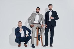 Three handsome young men in suits and bowties looking at camera while standing against gray background photo