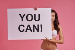 Encouraging others.  Attractive young woman covering a half of her face with poster and looking at camera while standing against pink background photo