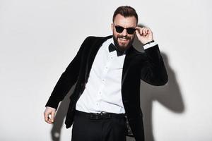 Man of charisma. Handsome young man in full suit adjusting his eyewear and smiling while standing against grey background photo