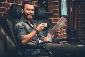 ahora este es un servicio apuesto joven barbudo mirando a la cámara con una sonrisa y sosteniendo una taza de café y periódico mientras se sienta en una silla cómoda en la barbería foto