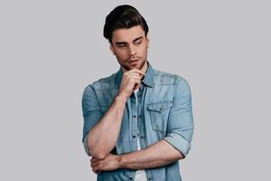 Thinking about... Good looking young man in casual wear keeping hand on chin and looking away while standing against grey background photo