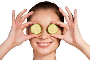 Cucumber spa. Beautiful young woman holding pieces of cucumber in front of her eyes and smiling while isolated on white background photo