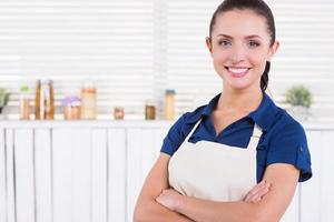 Ready to cook. Beautiful young woman in apron keeping arms crossed and smiling while standing in a kitchen photo