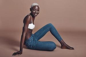 Feeling free to express herself. Attractive young African woman looking at camera and smiling while sitting against brown background photo