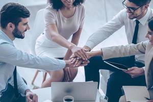 Ready for new achievements every day. Close-up part of top view of group of four young people holding hands and showing their unity with smile while sitting on the couch at office photo