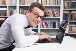 Computer geek. Side view of young nerd man in shirt and bow tie typing something on computer and looking at camera with smile photo