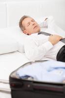 hombre de negocios cansado durmiendo. hombre cansado de pelo gris con camisa y corbata acostado en la cama y manteniendo los ojos cerrados mientras el equipaje está acostado en la cama foto