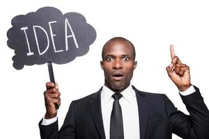 Just inspired. Surprised young African man in formalwear holding banner with text and pointing up while standing isolated on white background photo