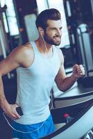 cinco millas con placer. un joven apuesto y alegre con auriculares mirando hacia otro lado con una sonrisa mientras corre en la cinta de correr en el gimnasio foto