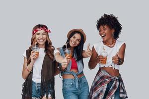 Three attractive young women keeping thumbs up and smiling while standing against grey background photo