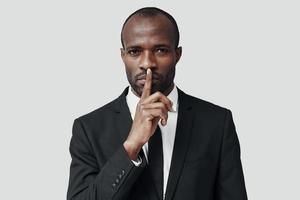 misterioso joven africano con ropa formal manteniendo el dedo en los labios mientras se enfrenta a un fondo gris foto