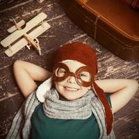 Happy little dreamer.Top view of happy little boy in pilot headwear and eyeglasses lying on the hardwood floor and smiling while wooden planer and briefcase laying near him photo