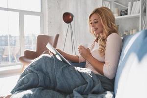 Interesting... Attractive young woman using digital tablet and smiling while sitting on the sofa at home photo