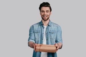 Gift for you Handsome young man in blue jeans shirt giving a gift box and looking at camera while standing against grey background photo