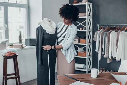 trabajando en un vestido nuevo. hermosa joven africana midiendo el vestido con cinta mientras está de pie en su taller con ropa colgada en el fondo foto