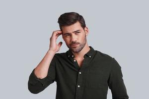 Feeling uncertain. Thoughtful young man touching his head with hand and looking away while standing against grey background photo